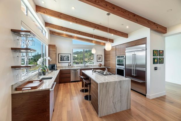 Bright, spacious contemporary kitchen with warm timber flooring, exposed timber ceiling beams, dark wood cabinetry, stone benchtops, stainless-steel appliances, and a large central island with seating. Glazed shelving and windows bring in natural light and views of greenery.