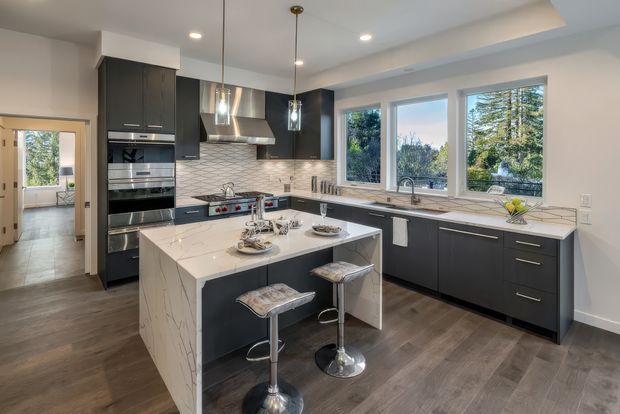 A spacious contemporary kitchen with a central island and two bar stools. Dark charcoal cabinetry contrasts with white marble-look benchtops and a white geometric tiled splashback. The kitchen includes stainless steel appliances, a gas cooktop and rangehood, pendant lights, under-cabinet lighting, and large windows overlooking greenery. Dark timber-look flooring and neutral walls complete the bright, polished space.