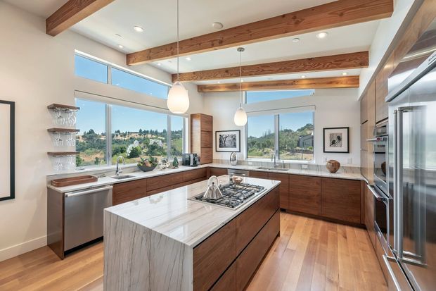 Bright, spacious contemporary kitchen with warm timber ceiling beams, timber flooring, dark wood cabinetry, and a large white veined stone island with integrated cooktop. Floor-to-ceiling glazing overlooks a green landscape, while stainless-steel appliances, open shelving, pendant lights, and recessed ceiling lights complete the space.