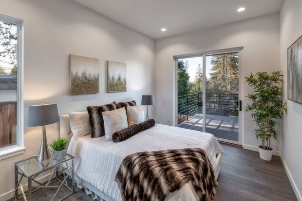 Bright, contemporary bedroom with a neatly made bed, layered neutral bedding and dark faux-fur throw, mirrored bedside table, table lamps, botanical artwork, indoor plants, recessed ceiling lighting, and dark timber-look flooring. Full-height glazed doors open onto a balcony with a black metal balustrade and leafy views.