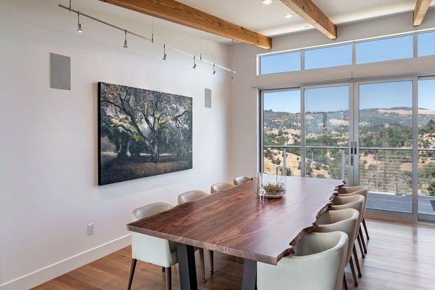 Bright contemporary dining room with a large live-edge timber dining table surrounded by upholstered chairs. White walls, warm timber flooring, exposed timber ceiling beams, track lighting, and full-height glazed doors and clerestory windows create an airy setting with expansive hillside views beyond.