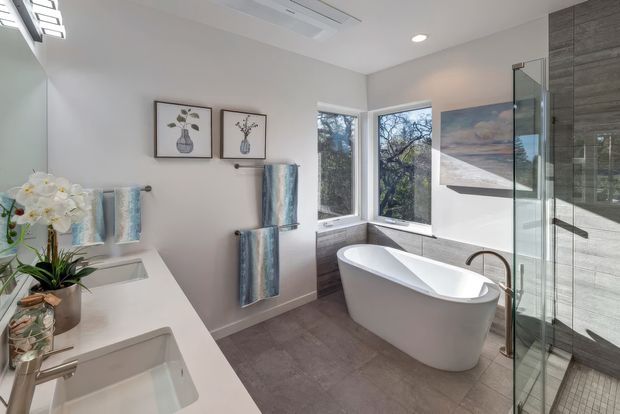 Bright, contemporary bathroom with a freestanding white bathtub positioned beside corner windows, a glass-enclosed shower, and a double vanity with white countertop and integrated basins. Grey tiled flooring and matching grey wall tiles in the shower area contrast with light painted walls. Brushed metal fixtures, patterned towels, framed botanical artwork, and a skylight-like ceiling opening add detail. Trees are visible through the windows.