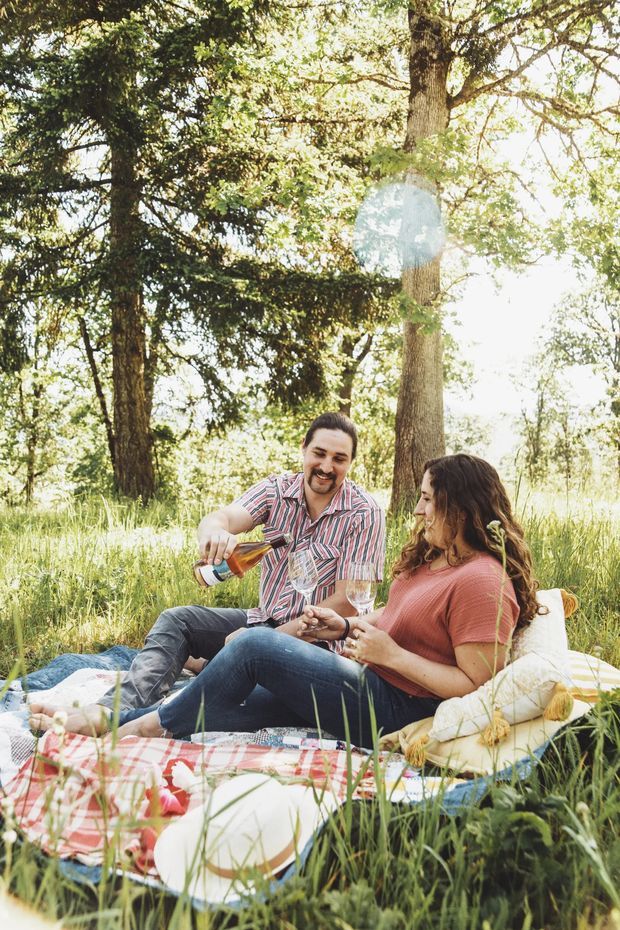 A sunlit outdoor lifestyle scene showing a couple relaxing on a picnic blanket in a grassy, wooded setting. They are seated among cushions and picnic accessories, sharing wine in a warm, natural environment. The image is a candid-style photograph with bright backlighting, tall trees, and lush vegetation.