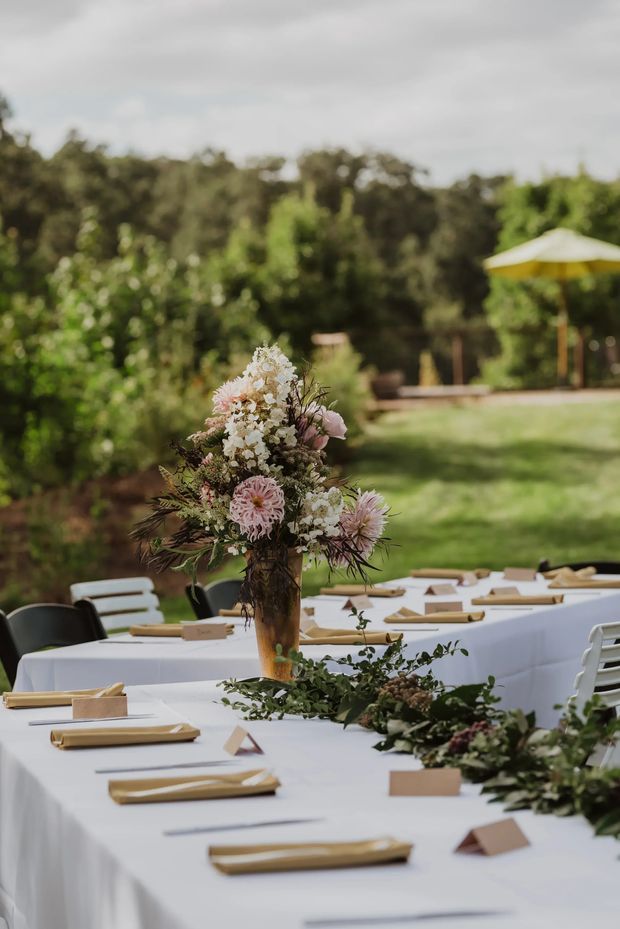 An outdoor garden dining setup with long tables covered in white linens, arranged place settings with tan napkins and cards, white and dark chairs, and lush floral and leafy centrepieces. The tables sit on a lawn surrounded by mature greenery, with a yellow-and-white parasol in the background. The image has a softly focused, warm, elegant event atmosphere.