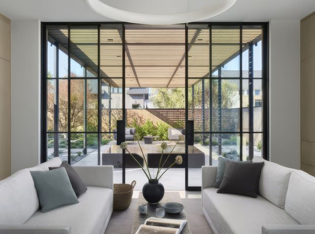 Bright contemporary living room with two white sofas facing each other around a low coffee table, opening through full-height black-framed glazing and doors to a covered outdoor terrace or courtyard. A large circular recessed ceiling feature, neutral finishes, and restrained furnishings create a refined indoor-outdoor setting.