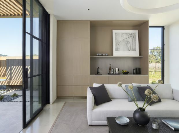 Bright contemporary living room with a large pale upholstered sofa, dark accent cushions, a black coffee table, and a full-height built-in timber-toned storage and display wall. Black-framed floor-to-ceiling glazing opens toward an outdoor courtyard, while a sculptural recessed ceiling detail and linear exterior shading add architectural interest.