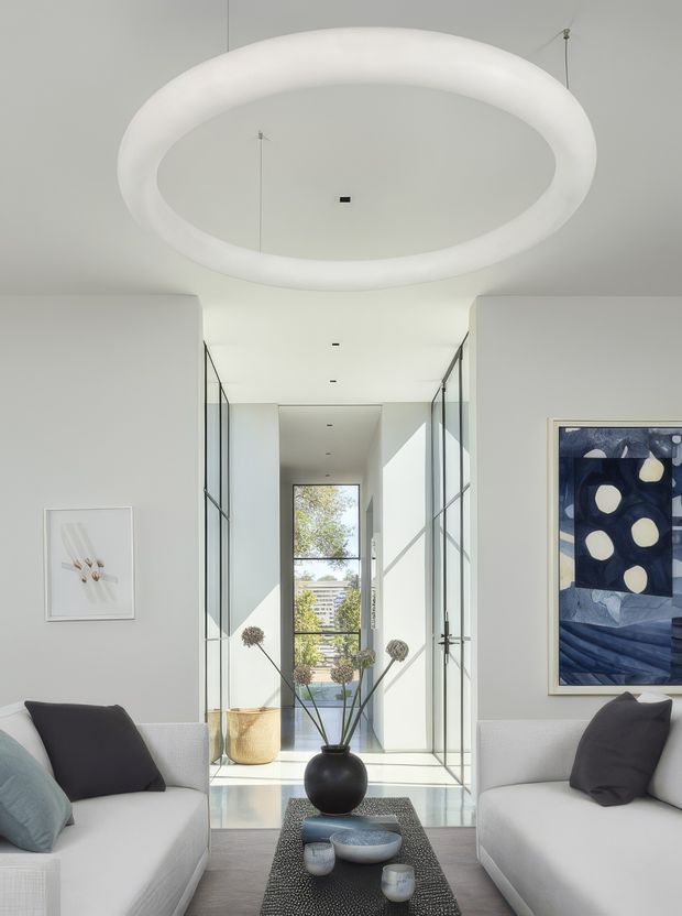 Bright, refined contemporary living room with two white sofas facing a narrow dark coffee table, arranged beneath a large suspended oval ring light. A glazed central hallway extends toward a rear window, creating strong natural light and visual depth. The room features pale grey walls, extensive glass partitions, minimalist recessed lighting, abstract artwork, and restrained black, white, and blue furnishings.