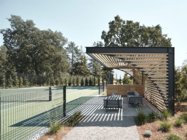 A contemporary outdoor pavilion beside a fenced tennis court, featuring a dark metal rectangular frame with a slatted timber pergola roof and privacy screen. A compact outdoor seating area sits on a paved terrace beneath the pergola, surrounded by landscaped grasses and mature trees. Strong sunlight creates striped shadows across the ground and furniture.