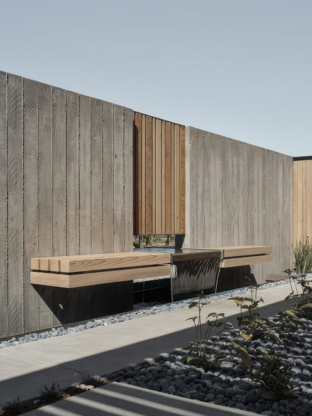 A minimalist outdoor courtyard or entry area featuring a floating timber bench and integrated dark basin or water feature against tall textured concrete walls. Vertical timber cladding and a narrow reflective panel add warmth and contrast. The ground is finished with smooth concrete paving and bands of dark rounded river stones with sparse planting. The composition is contemporary, sculptural, and strongly geometric.