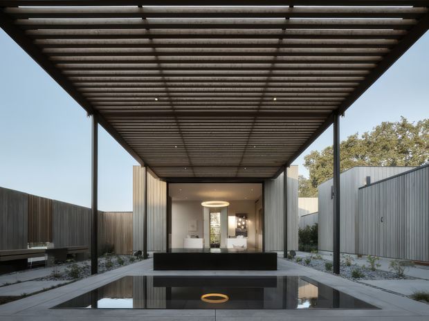 A contemporary covered courtyard or entry sequence framed by a large dark metal pergola with closely spaced slatted beams. A shallow reflecting pool occupies the foreground, aligned with a central path leading toward a glazed interior space. Corrugated metal-clad walls, dark timber fencing, pale gravel landscaping, and restrained planting create a minimalist, highly geometric composition. A circular pendant or reflected light feature provides a warm accent.