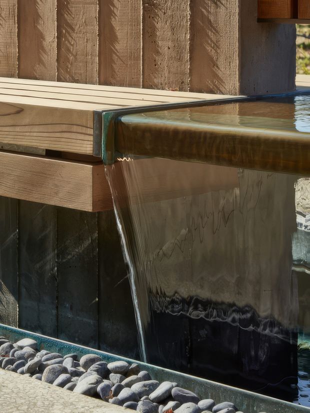 Close-up architectural detail of a pool or water feature with an engineered-looking sheet-like edge and flowing water spilling over a dark vertical wall. Timber-clad structure and a bed of smooth rounded stones frame the feature, with warm natural light and strong material contrast.